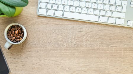 A modern workspace featuring a cup of coffee beans, keyboard, phone, and plant on wooden desk.